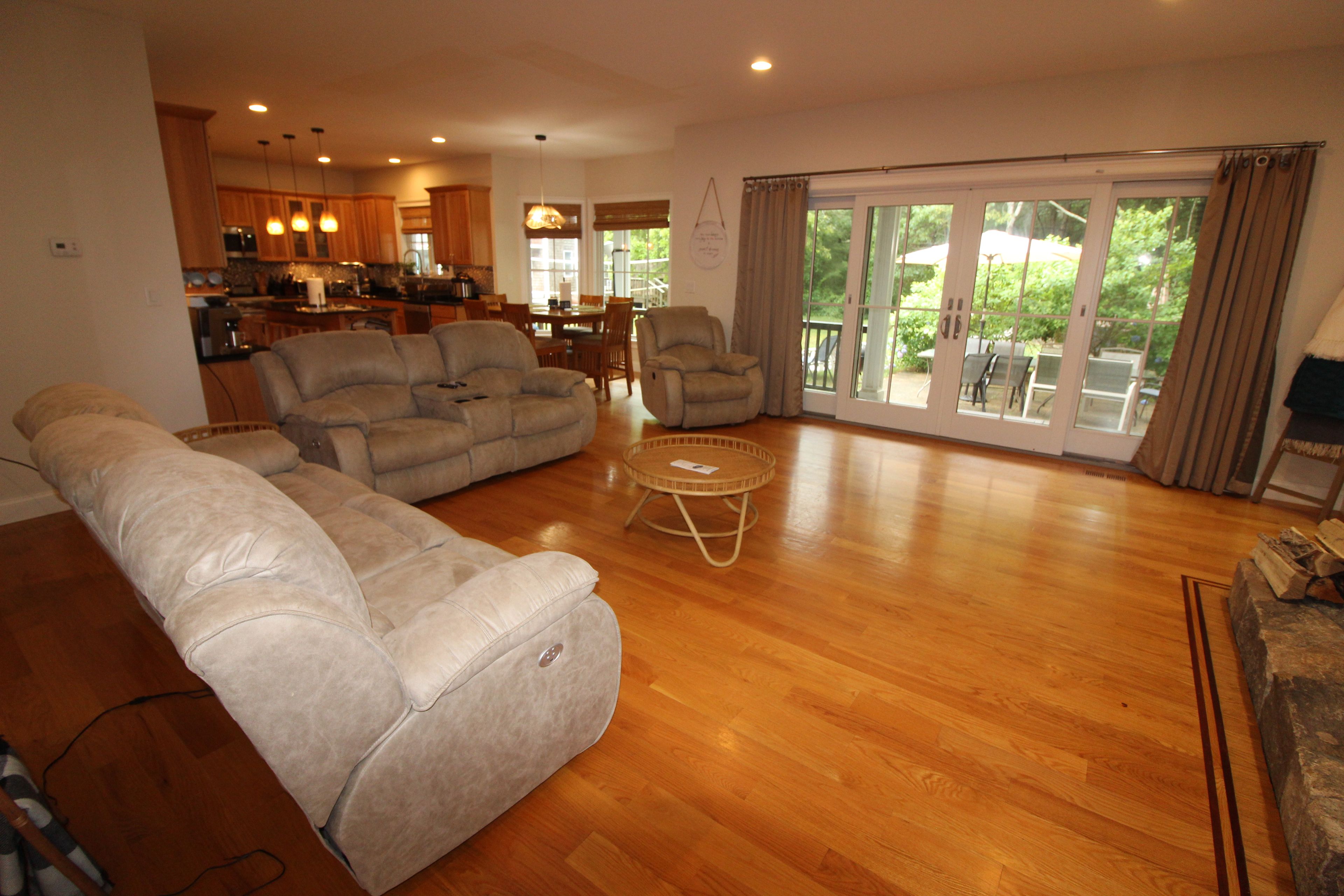 A spacious living room with a large sectional sofa, a smaller sofa, and a round coffee table. There is a dining area with a table and chairs, and a kitchen in the background.