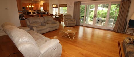 A spacious living room with a large sectional sofa, a smaller sofa, and a round coffee table. There is a dining area with a table and chairs, and a kitchen in the background.