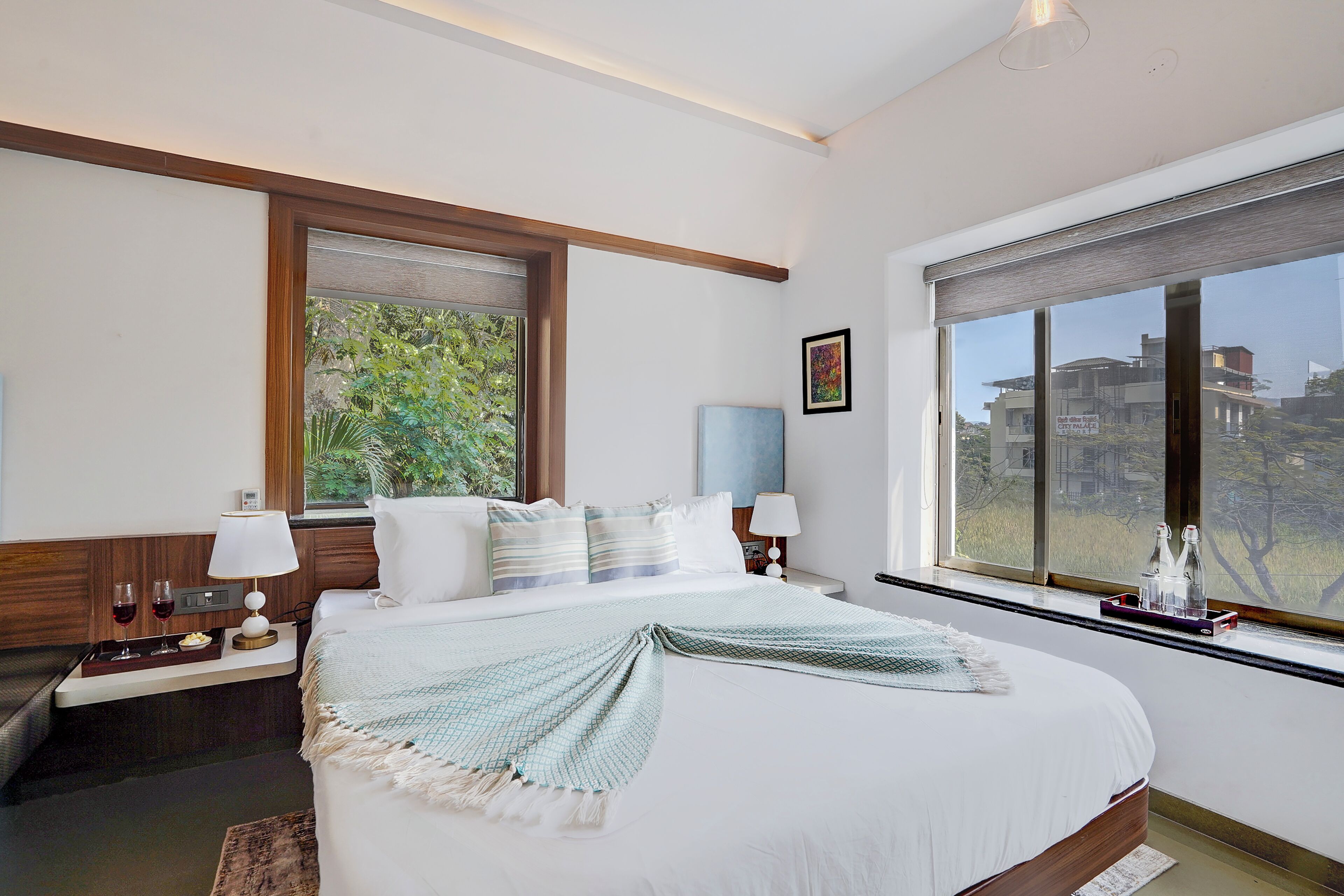 A hotel room with a large bed, a nightstand with a lamp, a window with a view of buildings, and a small table with a tray of items.