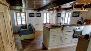 Een landelijke keuken met houten kasten, een gootsteen en een fornuis. In de aangrenzende kamer staat een bed met een geruit dekbed en kussens.