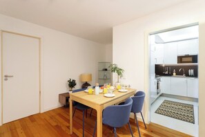 Eine kompakte Küche mit modernen Geräten, ein Essbereich mit einem Holztisch und blauen Stühlen sowie ein kleiner Beistelltisch mit Lampe und Topfpflanze.