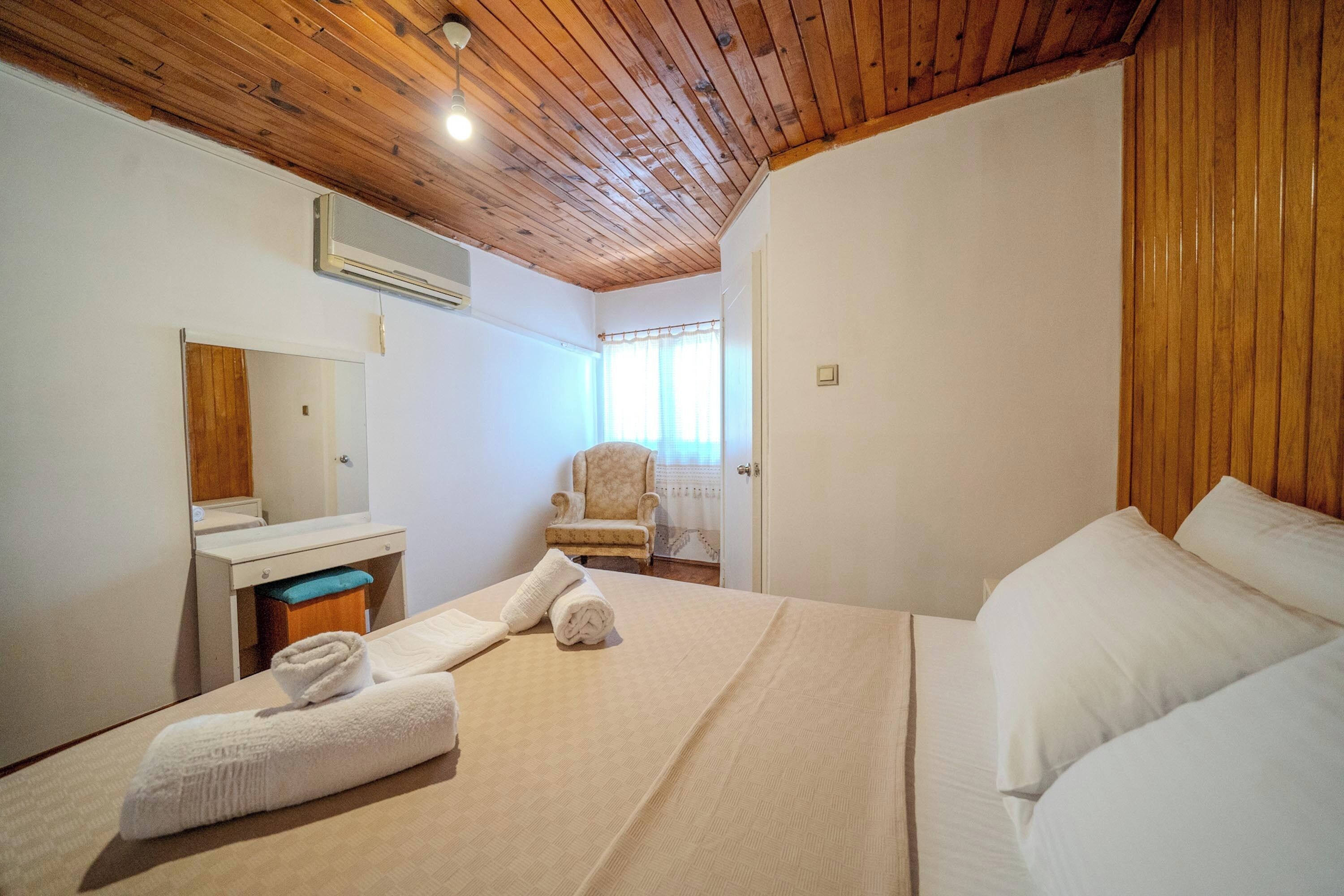 Una camera d'albergo con soffitto in legno, un letto con lenzuola bianche, una sedia, un tavolino con uno specchio e una finestra con tende.