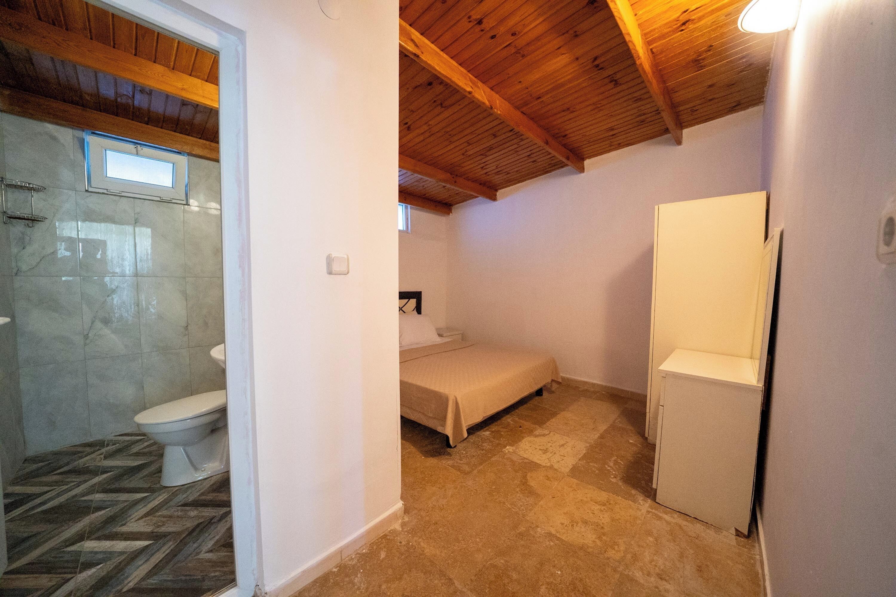 Una habitación pequeña y acogedora con techo de madera, cama, armario blanco y baño con inodoro y ducha.