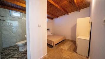Una habitación pequeña y acogedora con techo de madera, cama, armario blanco y baño con inodoro y ducha.