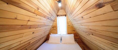 Una habitación pequeña de madera con techo inclinado, una cama con ropa blanca y una única lámpara.