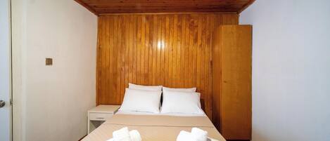 Habitación de hotel pequeña y con un solo ambiente, con techo de madera y una cama con ropa blanca. Hay toallas sobre la cama y una mesita de noche.