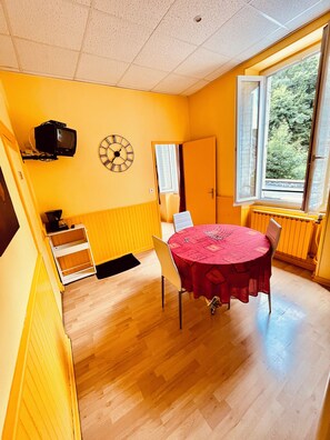A room with a wooden floor, a round table with a red tablecloth, a wall clock, a television mounted on the wall, and a window with a view of trees. - F1 30M2 2/4 persons (La Bourboule)