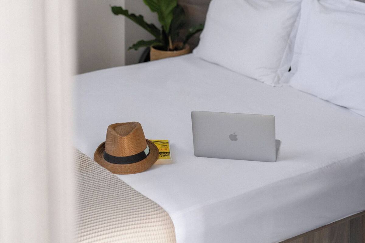 A neatly made bed with white linens, a laptop, a straw hat, and a potted plant on the nightstand.