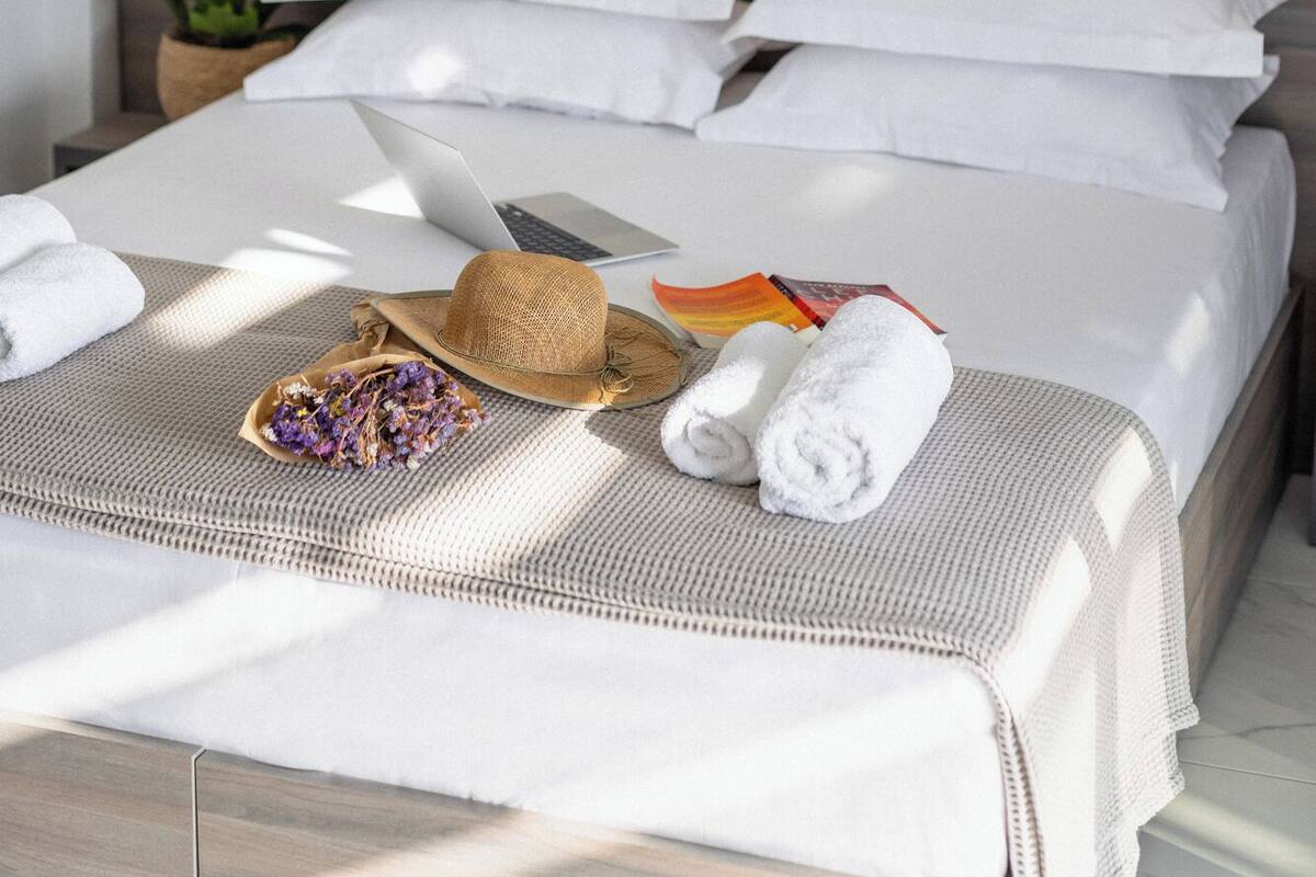 A neatly made bed with white linens, a laptop, a straw hat, and a bouquet of dried flowers on the bed.