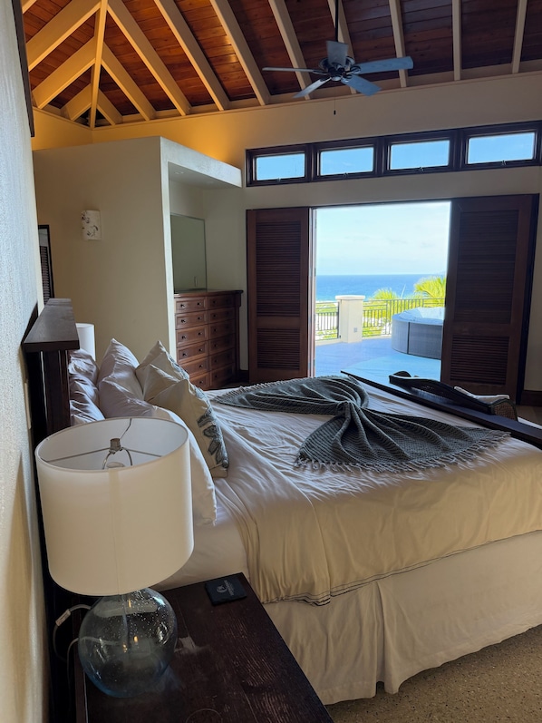 A bedroom with a bed, a nightstand, a lamp, a ceiling fan, and a view of the ocean through large windows.