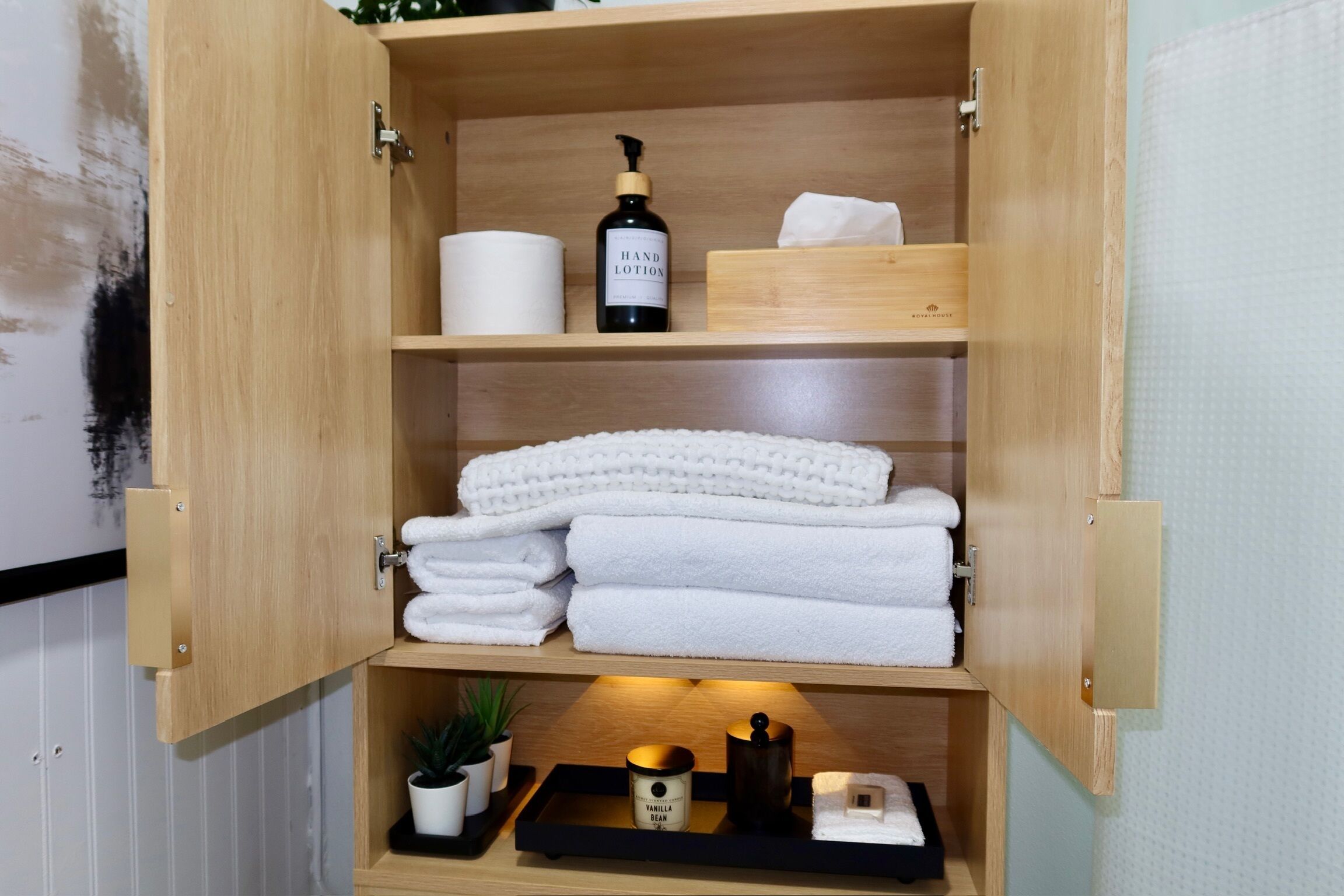 A wooden cabinet with shelves containing white towels, a hand lotion bottle, a candle, and a small plant.