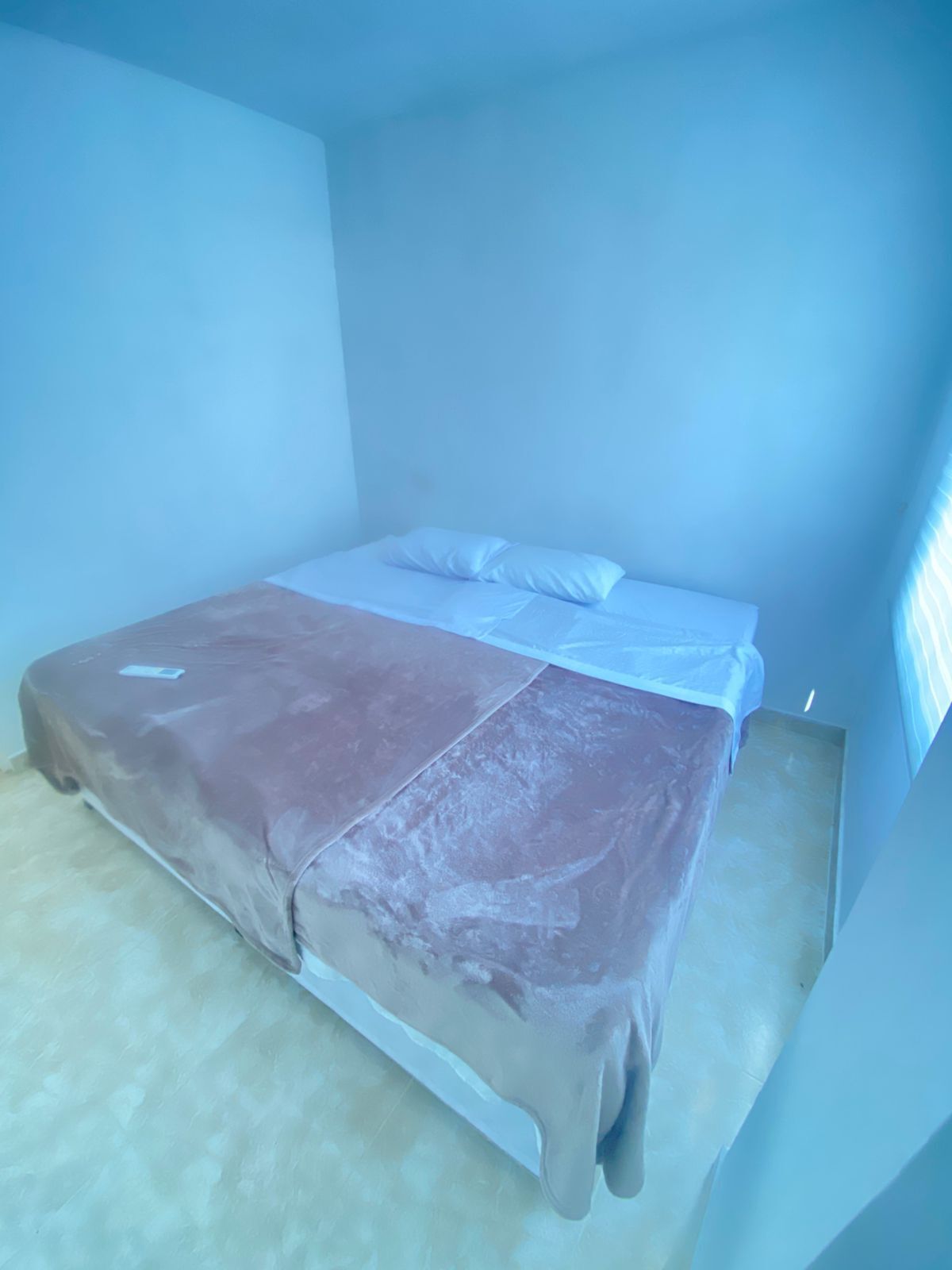 Una cama individual con colchón y almohadas blancos, una manta marrón y una alfombra de color claro en una habitación con paredes lisas y techo inclinado.