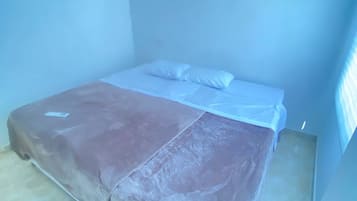 Una cama individual con colchón y almohadas blancos, una manta marrón y una alfombra de color claro en una habitación con paredes lisas y techo inclinado.