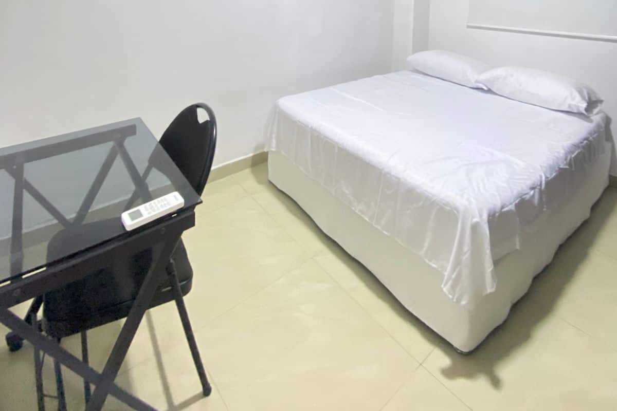 A small, clean room with a bed, a desk, and an air conditioning unit.