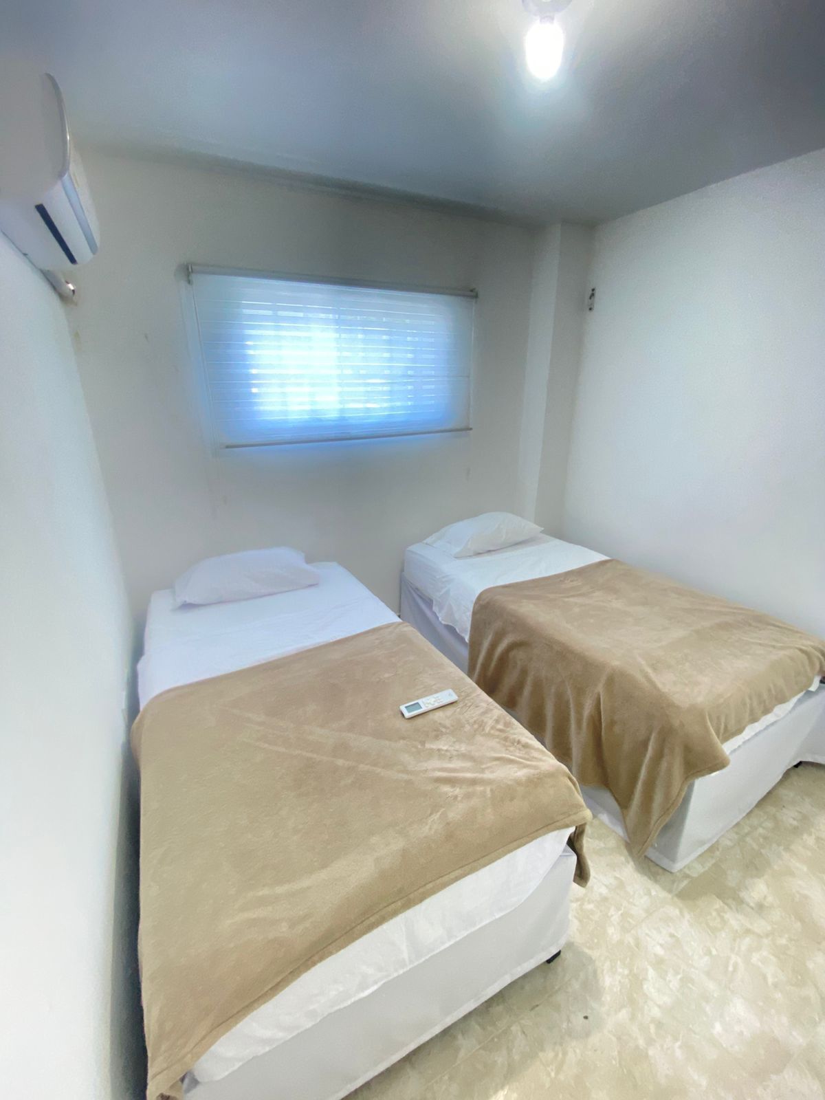A room with two beds, a window with blinds, a thermostat, and a light fixture.