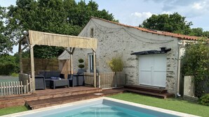 Une piscine avec une terrasse en bois et des meubles d’extérieur, un bâtiment en pierre avec un toit en tuiles et une porte blanche.