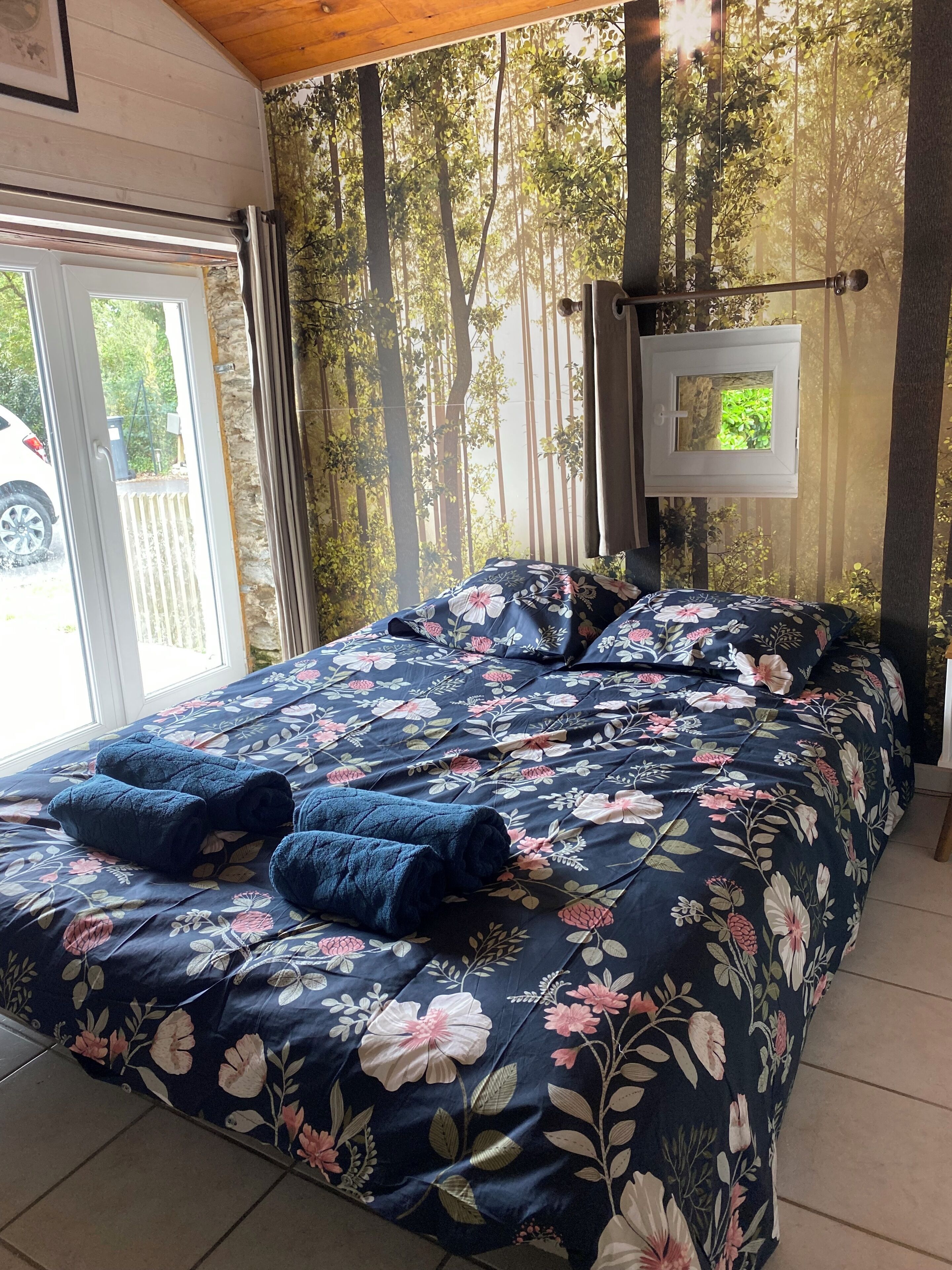 A bedroom with a floral bedspread, a view of trees through a window, and a door leading outside.