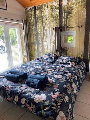 Ein Schlafzimmer mit floralem Bettüberwurf, Blick auf Bäume durch ein Fenster und einer zur Außenwelt führenden Tür.