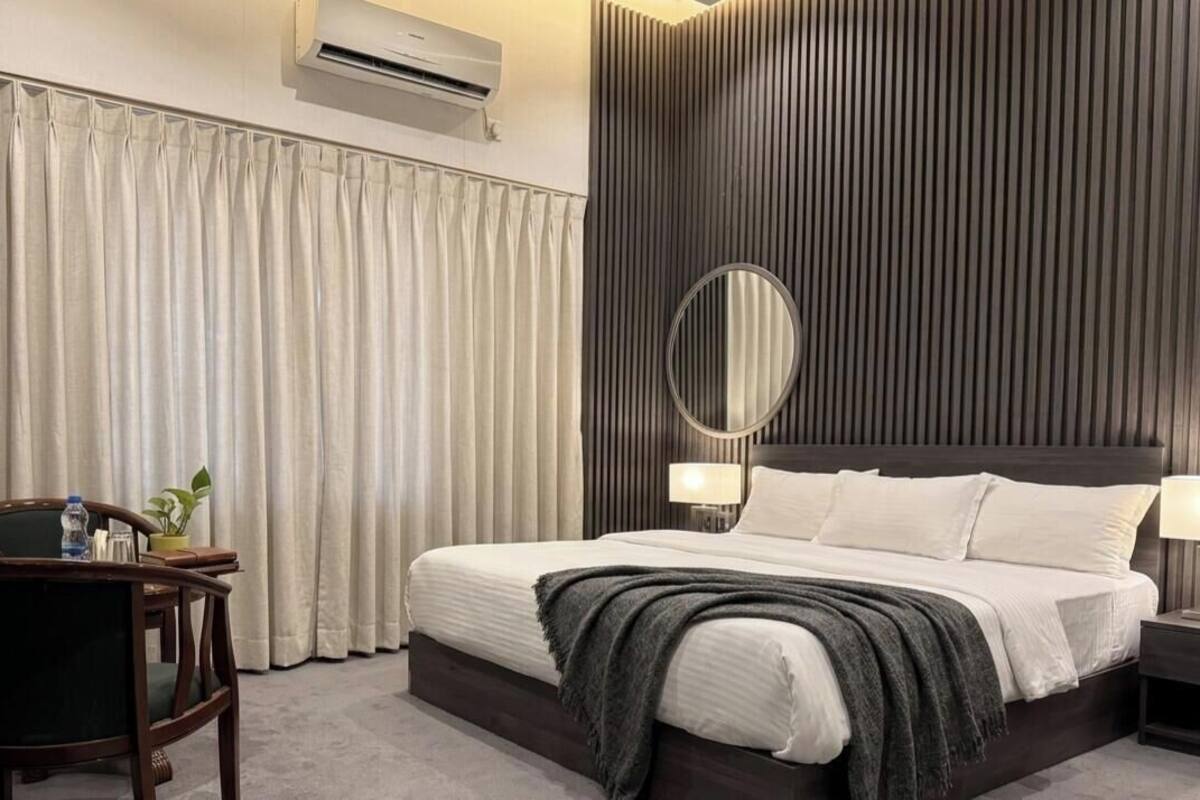 Habitación de hotel con cama, una silla, mesita de noche con lámpara, espejo y ventana con cortinas.