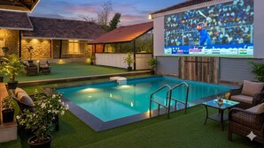 Una piscina con una pantalla de proyección mostrando un partido de cricket, rodeada de mobiliario de exterior y plantas.