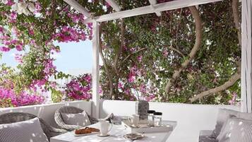 Uma área de estar ao ar livre com mobiliário branco, uma mesa posta com xícara, pires e um prato de comida, cercada por flores cor-de-rosa em flor.