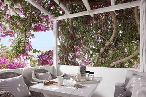 Uma área de estar ao ar livre com mobiliário branco, uma mesa posta com xícara, pires e um prato de comida, cercada por flores cor-de-rosa em flor.
