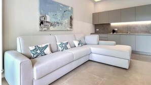 A modern living room with a white sectional sofa, a large abstract painting, and a kitchen area with grey cabinets. - Hostdomus - Princess Suite (Marina di Andora)