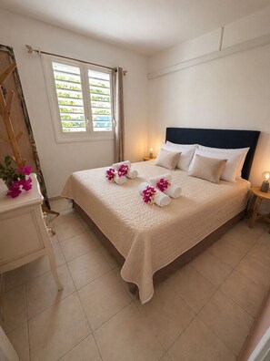 A neatly made bed with a quilted cover, a bedside table with a lamp, and a window with curtains. - Cosy sweet Home - Villa 4-6 pers - Swimming pool and Quiet area (Sainte-Anne)