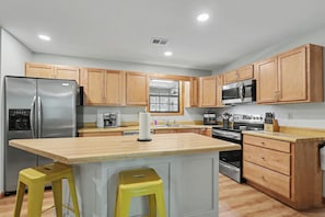 A kitchen with wooden cabinets, stainless steel appliances, a central island with stools, and a window above the sink. - Refresh and Renew at Margaret’s Place on Lake Chatuge! (Hiawassee)