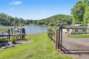 A lakeside view with a fenced area, a building with a green roof, and a grassy area with outdoor seating. - Refresh and Renew at Margaret’s Place on Lake Chatuge! (Hiawassee)
