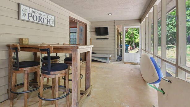 Eine Veranda mit Schild, Holzstühlen, einem Tisch und einem an der Wand befestigten Fernseher.