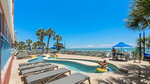 A pool area with lounge chairs, a small pool, and palm trees. - Oceanfront 2BR 2BA, Pools, Hot Tub, Epic Views (Myrtle Beach)