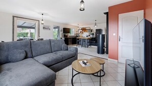 A modern living room with a grey sofa, two round wooden tables, a TV, and a kitchen in the background. - Villa Joël – House with Private Pool, 10 Minutes from the Beach (La Jonchère)