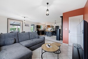 A modern living room with a grey sofa, two round wooden tables, a TV, and a kitchen in the background.