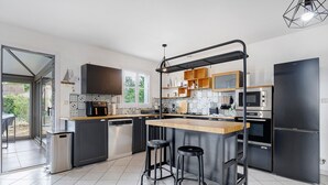 A modern kitchen with a central island, bar stools, built-in appliances, and a view through the glass door. - Villa Joël – House with Private Pool, 10 Minutes from the Beach (La Jonchère)