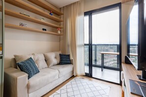Moderni olohuone, jossa on sohva, taulutelevisio, kirjoja sisältävä puinen hylly ja suuri ikkuna, josta on kaunis näkymä.