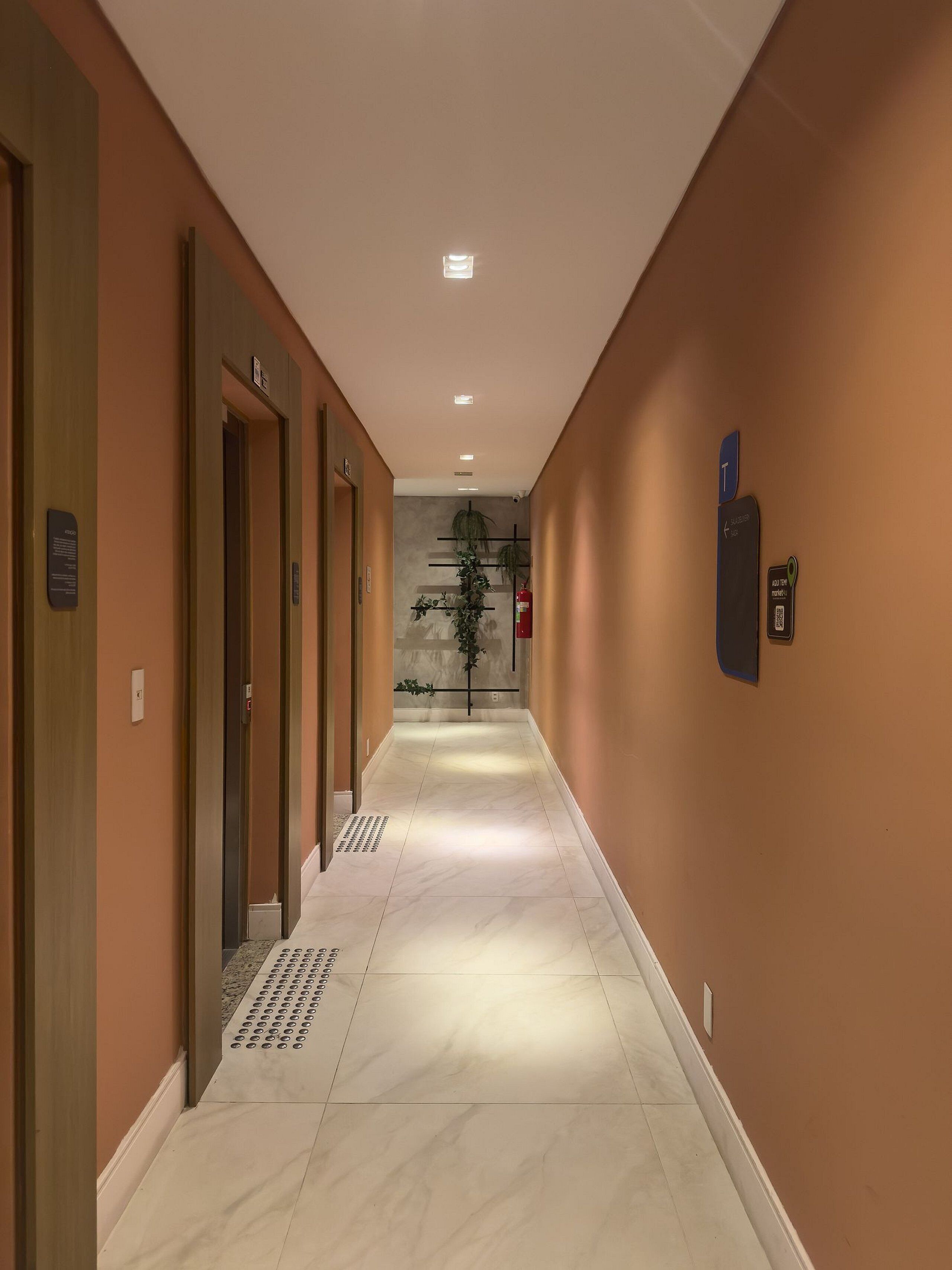 A hotel hallway with multiple doors, a fire extinguisher, a QR code, and a plant.
