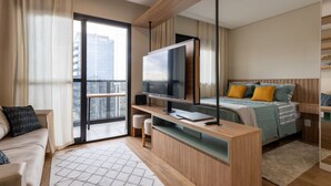 A modern bedroom with a large TV, a bed with patterned bedding, a sofa, and a large window. - Studio recém-construído no coração da Vila Olímpia (São Paulo)