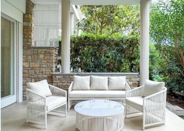Un divano di rattan bianco con cuscini, un tavolino da caffè in vetro tondo e un muro di pietra sullo sfondo.