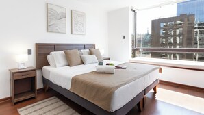 Ein modernes Schlafzimmer mit einem großen Bett, einem Nachttisch, zwei Wandbildern und einem Fenster mit Blick auf Gebäude.