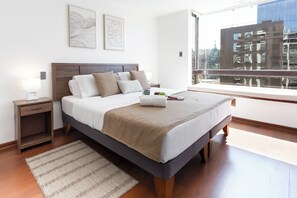 Ein modernes Schlafzimmer mit einem großen Bett, einem Nachttisch, zwei Wandbildern und einem Fenster mit Blick auf Gebäude.