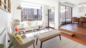 Ein modernes Wohnzimmer mit einer Couch, einer Bank und einem Essbereich, große Fenster mit Blick auf die Stadt und eine Treppe, die zu einer oberen Etage führt.