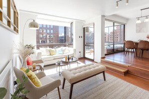 Ein modernes Wohnzimmer mit einer Couch, einer Bank und einem Essbereich, große Fenster mit Blick auf die Stadt und eine Treppe, die zu einer oberen Etage führt.