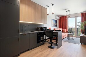 A modern kitchen with dark cabinetry, a built-in oven, and a dining area with a bar stool and a sofa. - Modern Apartments with Balconies | Downtown (Gdańsk)