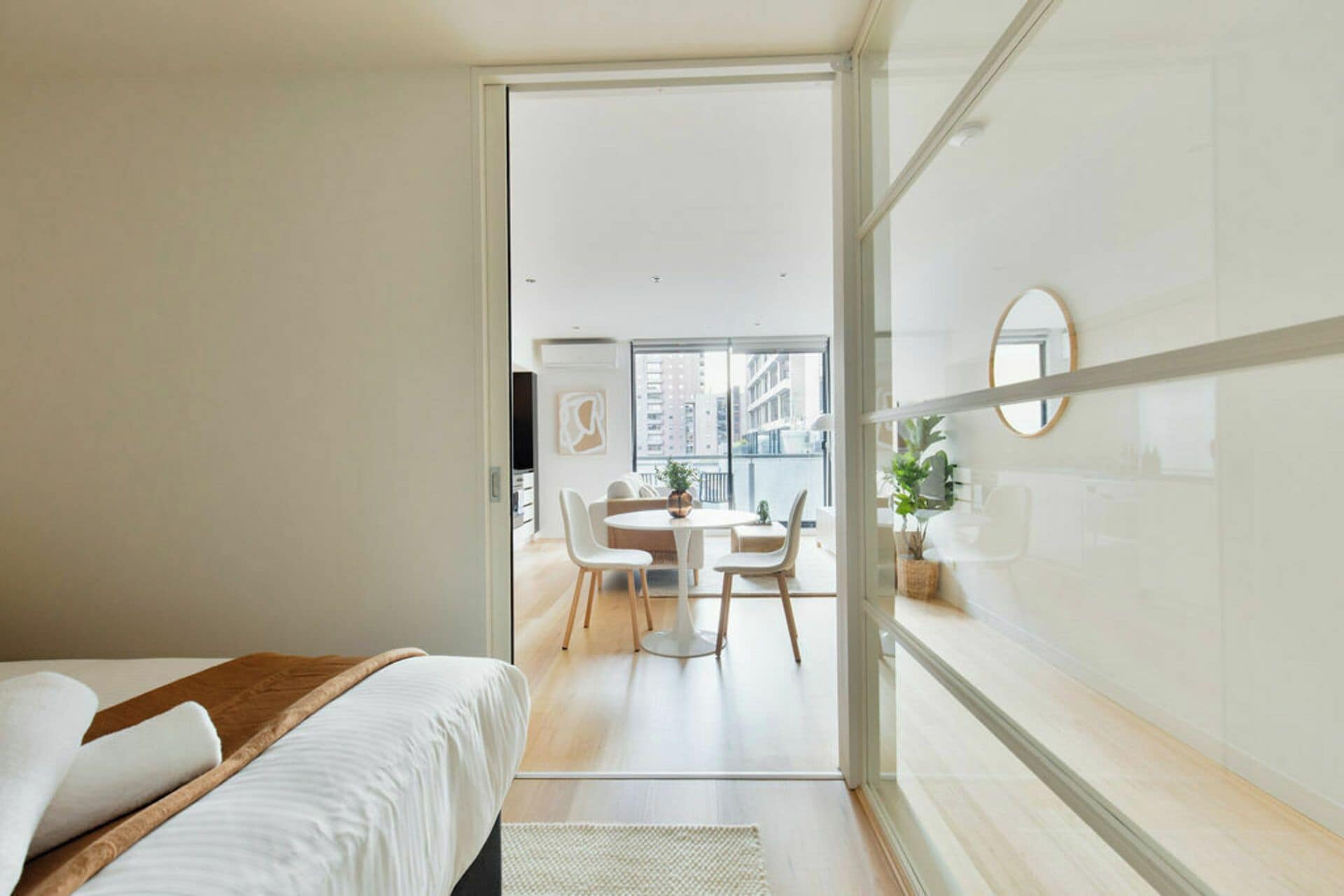 Une chambre d’hôtel moderne comprenant un lit, une cloison en verre, un coin repas avec des chaises et une table, et une vue sur la ville grâce à de grandes fenêtres.