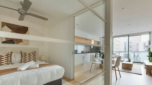 Een moderne hotelkamer met een bed, een plafondventilator, een kitchenette en een balkon met zitplaatsen.