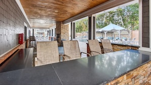 A covered outdoor seating area with a stone wall, wooden ceiling, and large windows overlooking a pool. - Landing Furnished Apartments Ocean Springs - Gulf Hills (Ocean Springs)