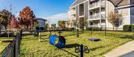 Un espacio al aire libre con césped, un tobogán azul, un parque infantil vallado y un edificio con balcones.