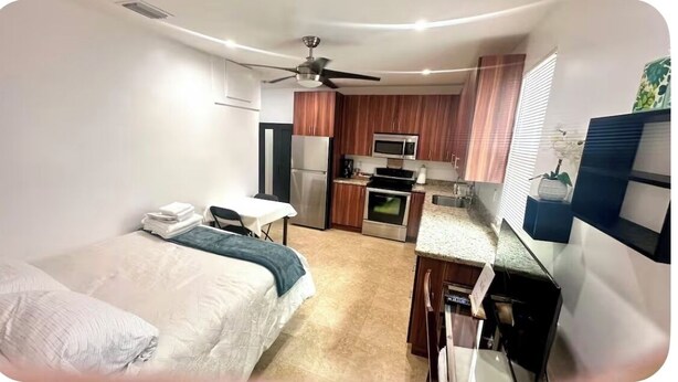 Een kleine kamer met een bed, bureau, kitchenette en plafondventilator.
