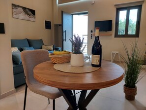 A dining area with a round wooden table, a wicker basket, a potted plant, and a wine bottle. - Elea Suites - Olive - Adelianos Kampos (Adelianos Kampos)